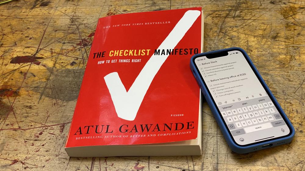 The book The Checklist Manifesto lays on a desk next to a phone with a checklist displaying.